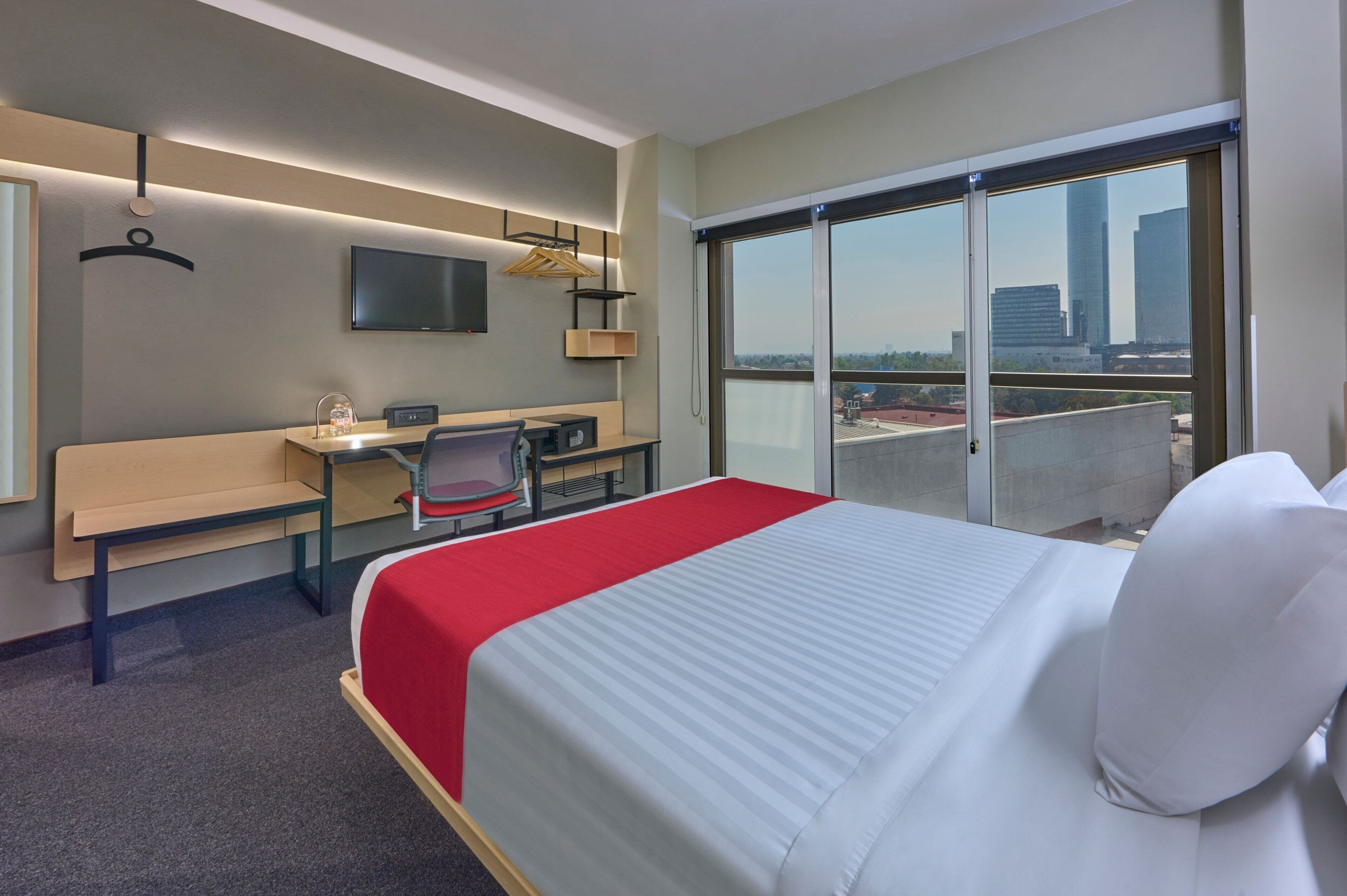 Quarto de hotel com cama, escrivaninha, cadeira, televisão e uma grande janela com vista para a cidade.