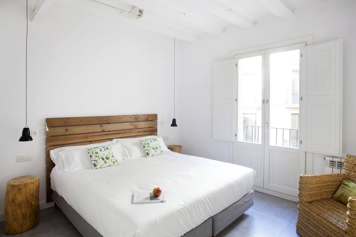 Quarto com cabeceira de madeira, roupa de cama branca, um banquinho de madeira pequeno e uma cadeira de vime.