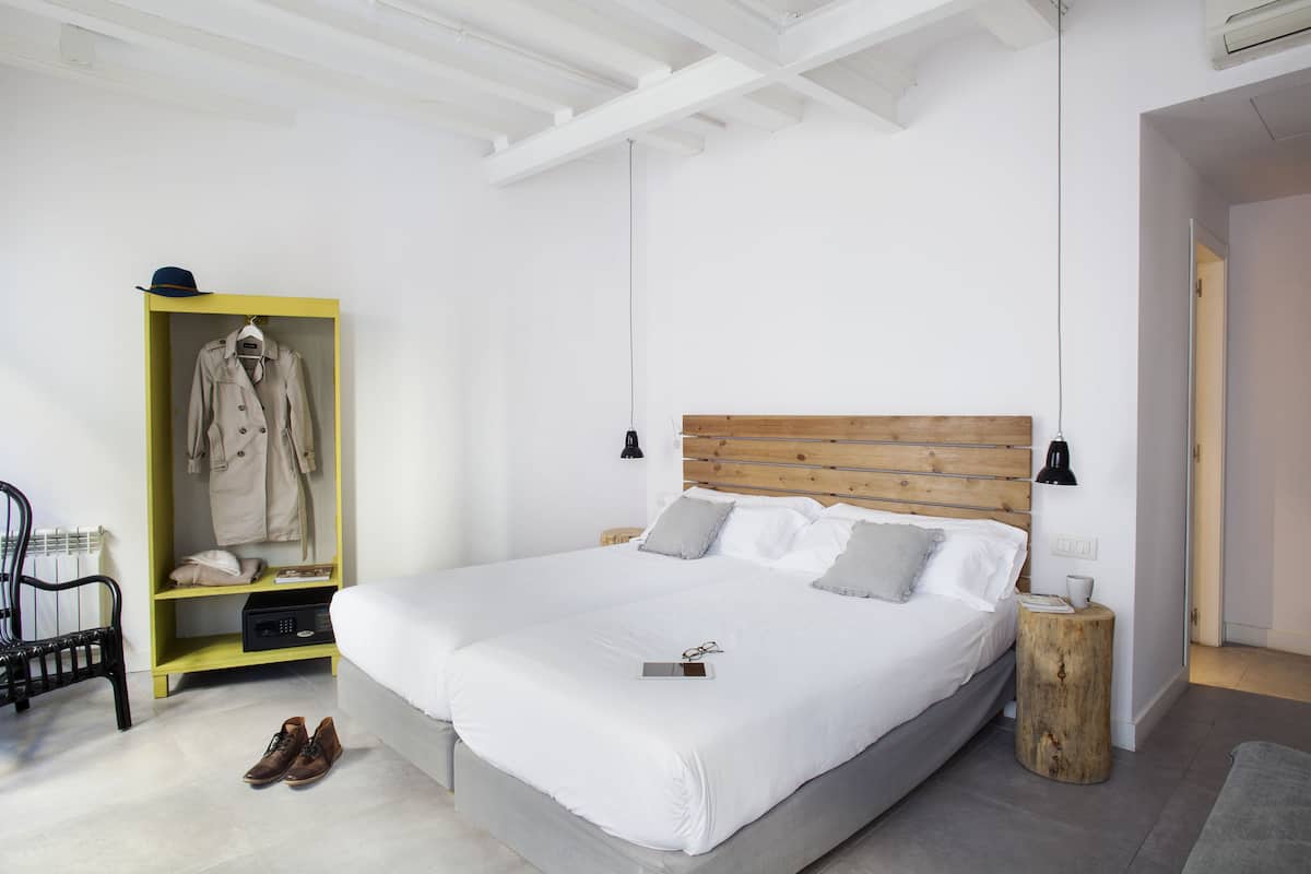Quarto arrumado com cama grande, cabeceira de madeira, luminárias de cabeceira, um banquinho de madeira pequeno, um cabideiro com um casaco e um par de sapatos no chão.