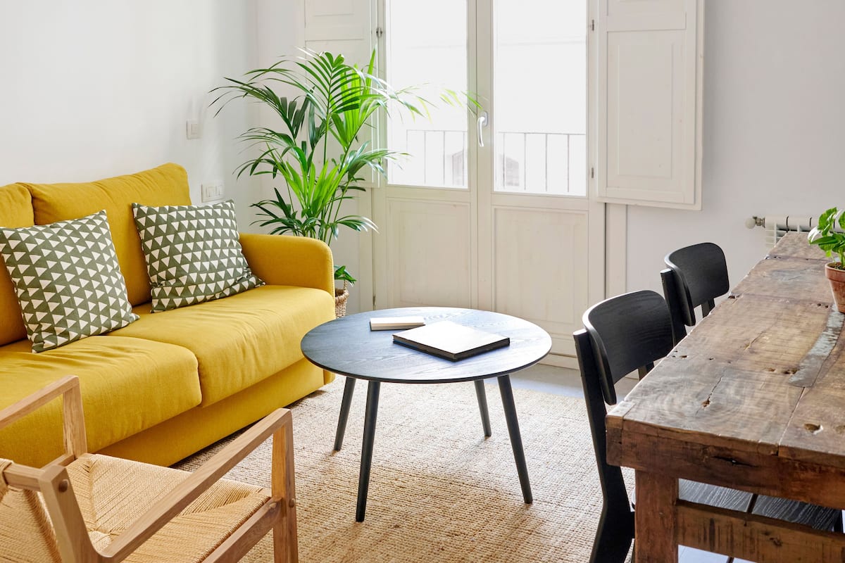 Uma sala de estar com um sofá amarelo, uma mesa de madeira redonda e um vaso com planta.