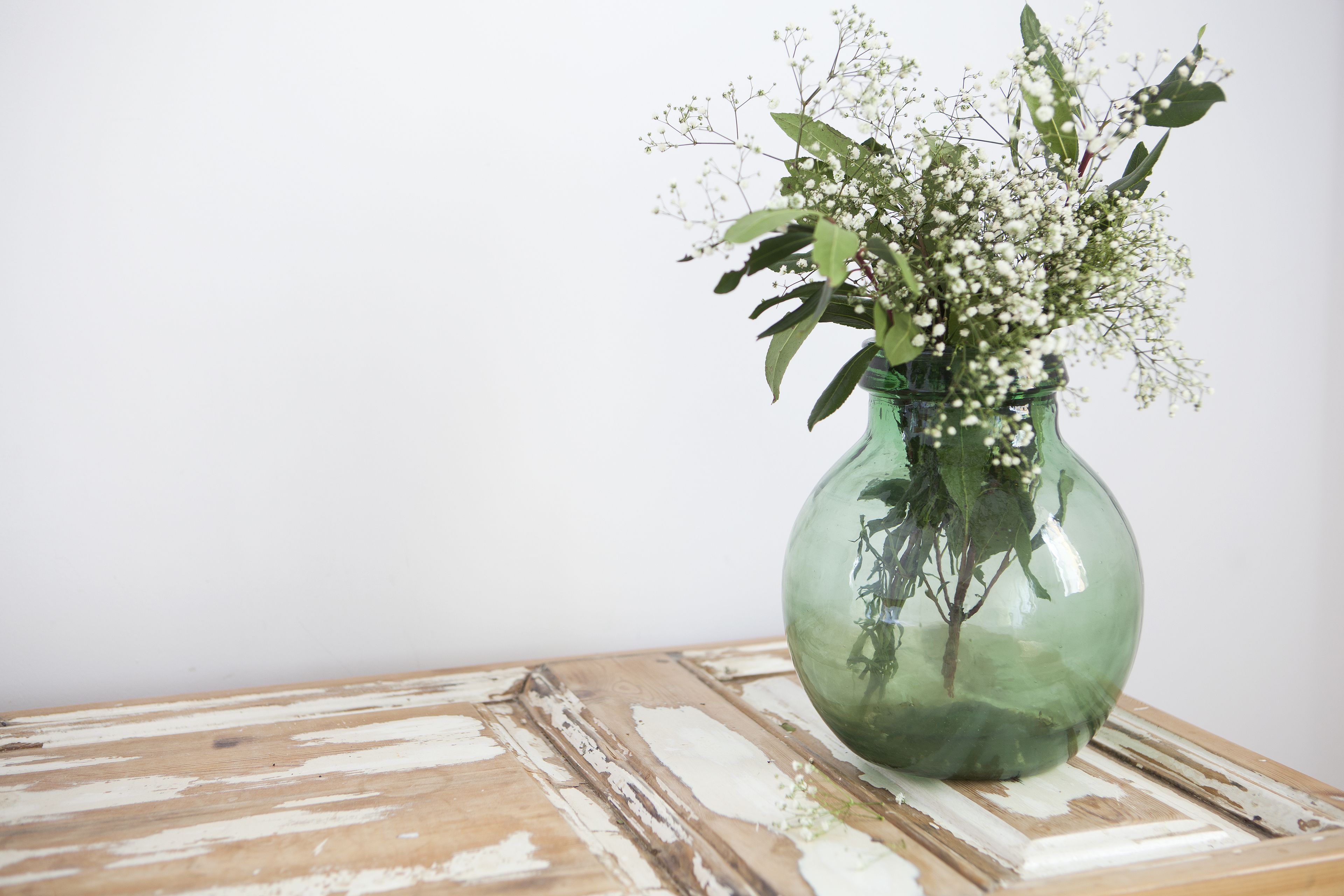 Un jarrón verde de vidrio con flores blancas y hojas verdes sobre una mesa de madera.