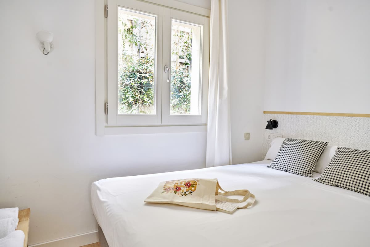 Cama bem arrumada com almofadas xadrez, janela com vista para o verde e parede branca com luminária.