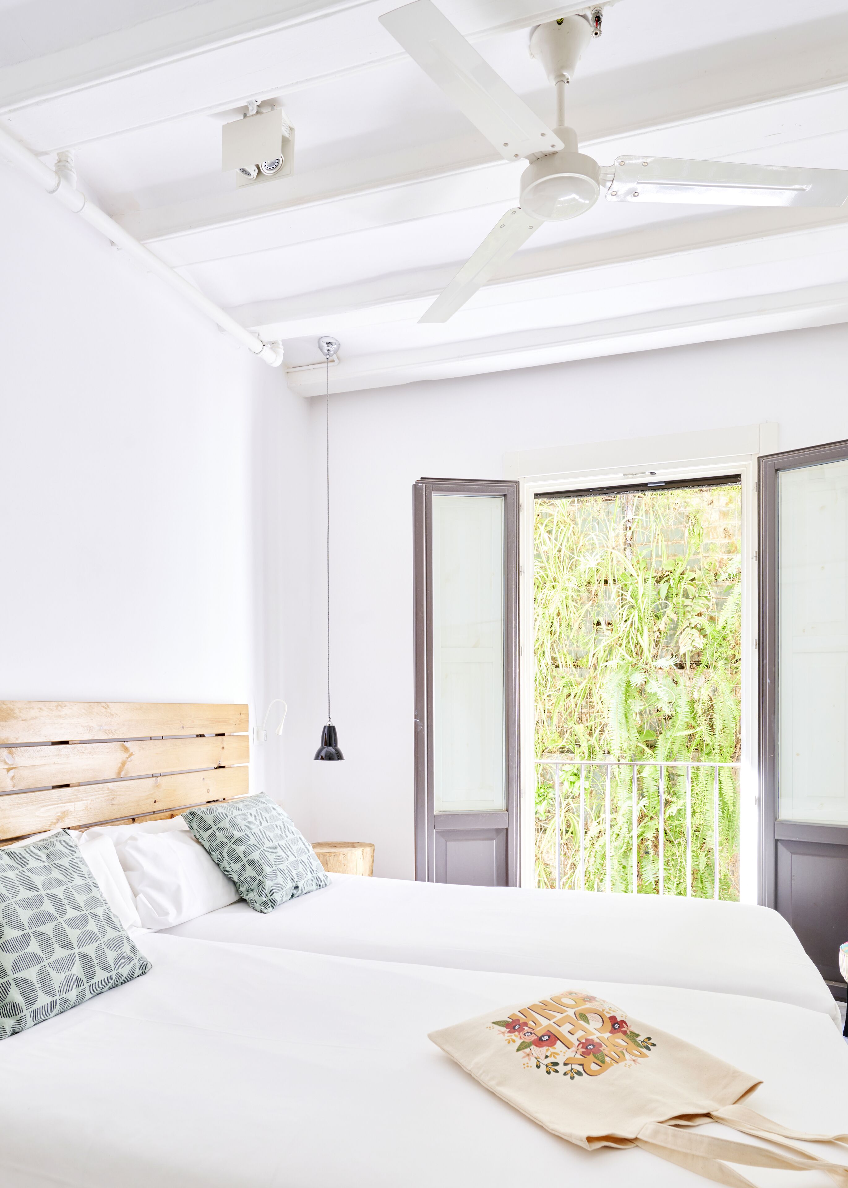 Un dormitorio con cabecera de madera, ropa de cama blanca, ventilador de techo y vistas a la vegetación a través de una ventana.