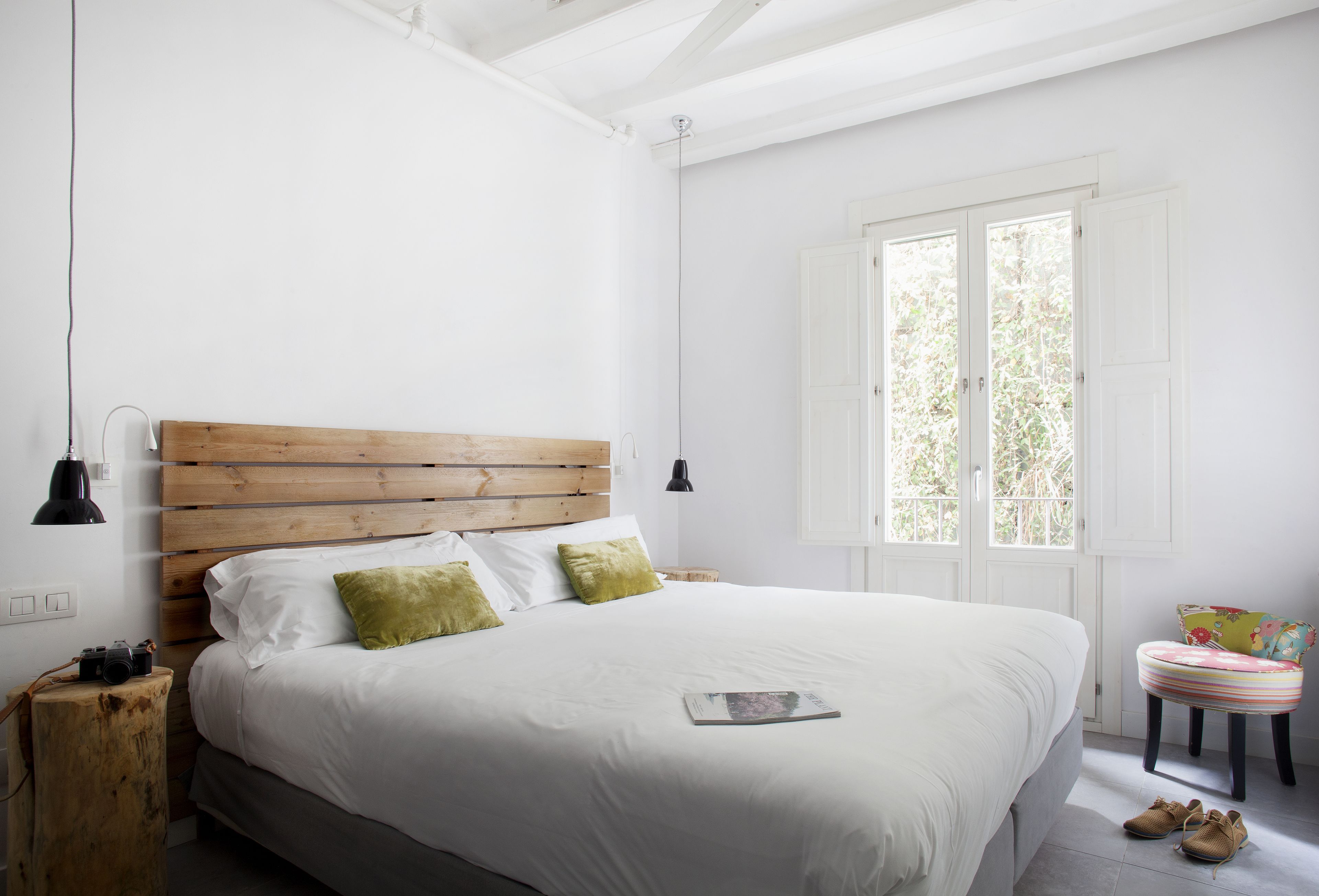 A bedroom with a wooden headboard, white bedding, a small wooden stool, and a pair of shoes on the floor.
