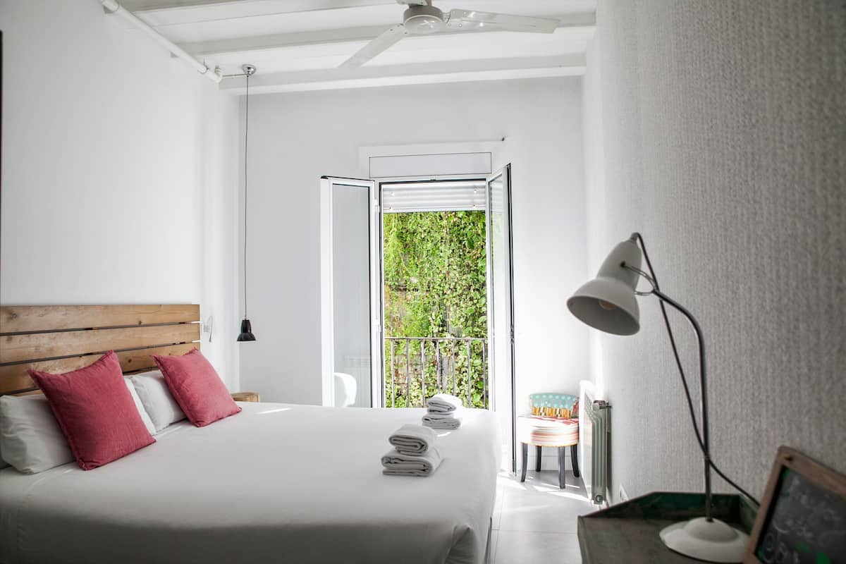 Quarto com cama, almofadas, mesa de cabeceira, abajur e vista para o verde através de uma porta aberta.