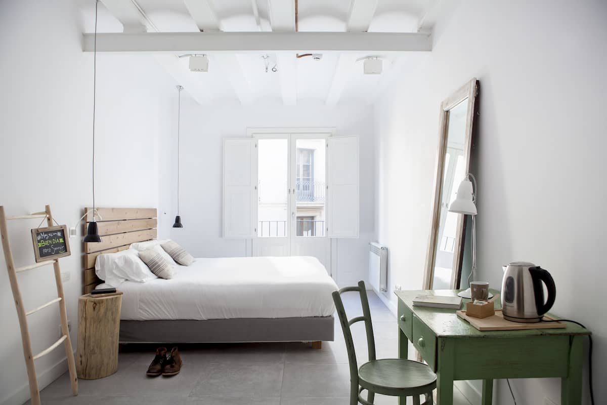 Quarto minimalista com cama, mesa de cabeceira de madeira, uma cadeira, escrivaninha com chaleira e janela com vista.
