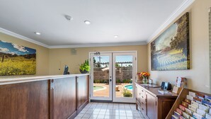 A reception area with a wooden counter, a large painting, a glass door leading outside, and a bookshelf with books. -  GoodSpring Motel (Napa)