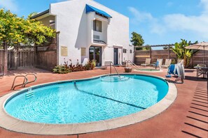 A swimming pool with a wooden deck, a white building with a blue awning, and a fenced area with lounge chairs.