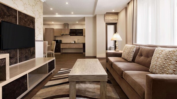 A modern living room with a flat-screen TV, a sofa, a coffee table, and a kitchen in the background. - Ramada by Wyndham Bishkek Centre (Bishkek)