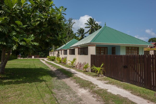 A bungalow with a green roof, surrounded by a wooden fence and lush greenery.