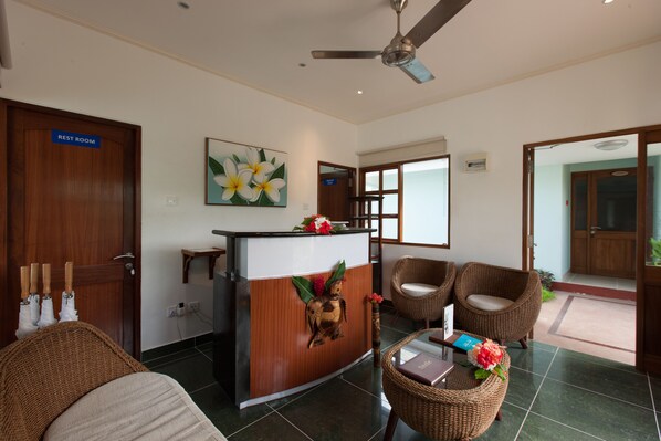 A reception area with a wooden desk, wicker chairs, a ceiling fan, and a painting of flowers. - Le Relax Self-Catering (La Digue)