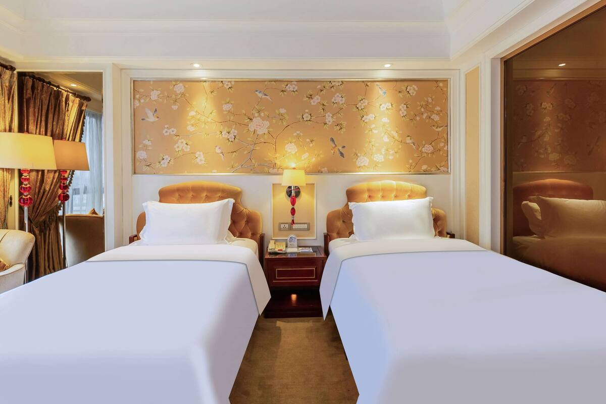A hotel room with two beds, a nightstand with a lamp, a floral wallpaper, and a view of the city.