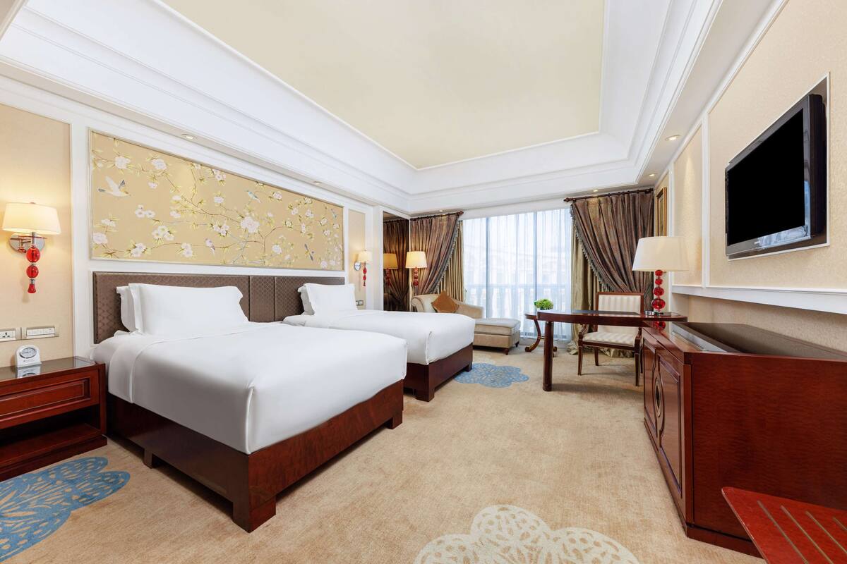 A hotel room with two beds, a TV, a desk, and a view of the city.