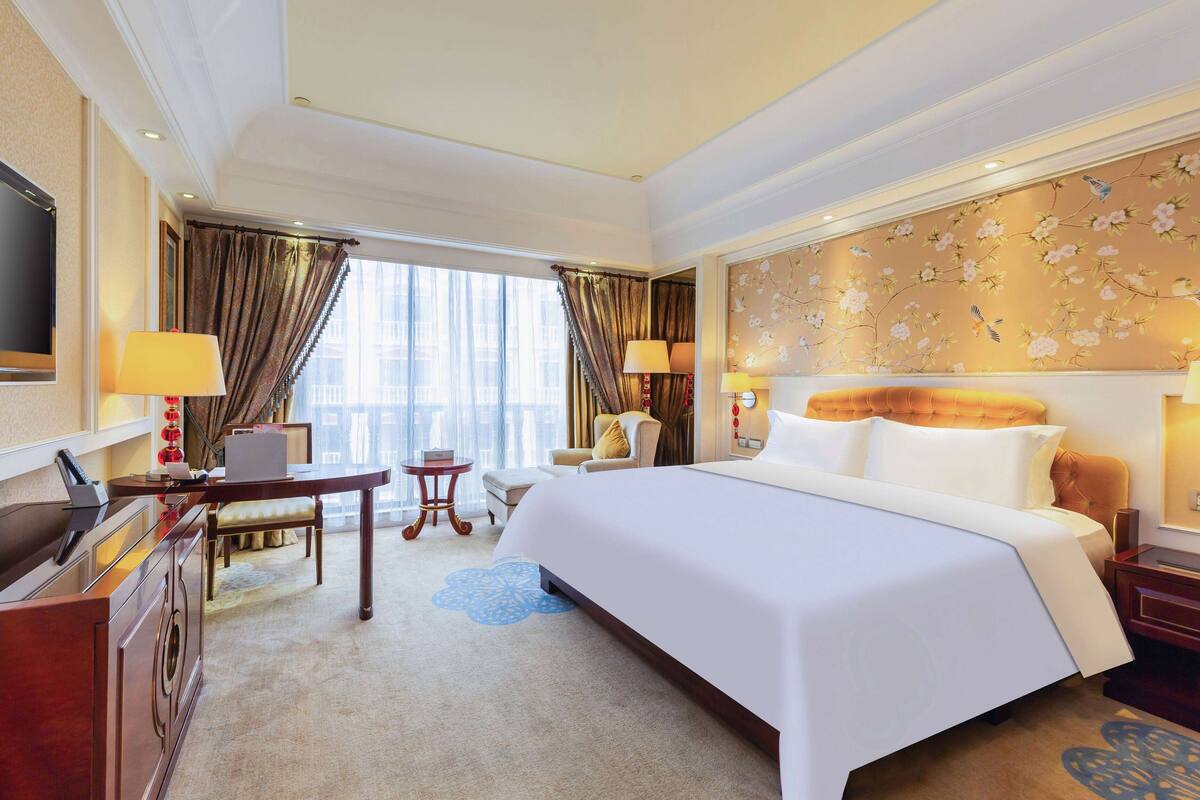 A hotel room with a large bed, a desk, a chair, a TV, and a view of the city.