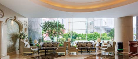 A spacious lobby with a marble floor, seating area with sofas, a reception desk, and a large window offering a view of the outdoors.