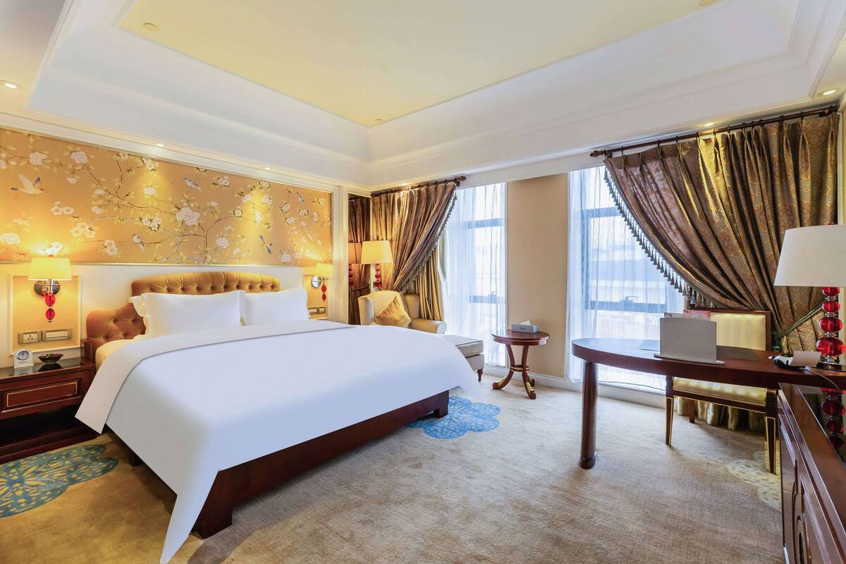 A hotel room with a large bed, a desk, and a chair. There is a window with curtains and a lamp on the desk.