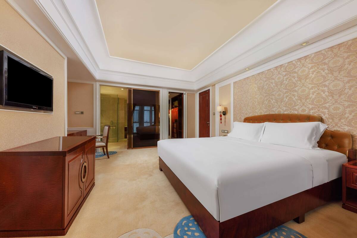 A hotel room with a large bed, a TV mounted on the wall, a wooden dresser, and a carpeted floor.