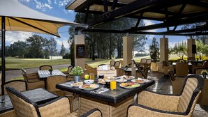 Um pátio externo com mobiliário de vime, uma mesa com comida e bebidas, e vista para um campo de golfe.