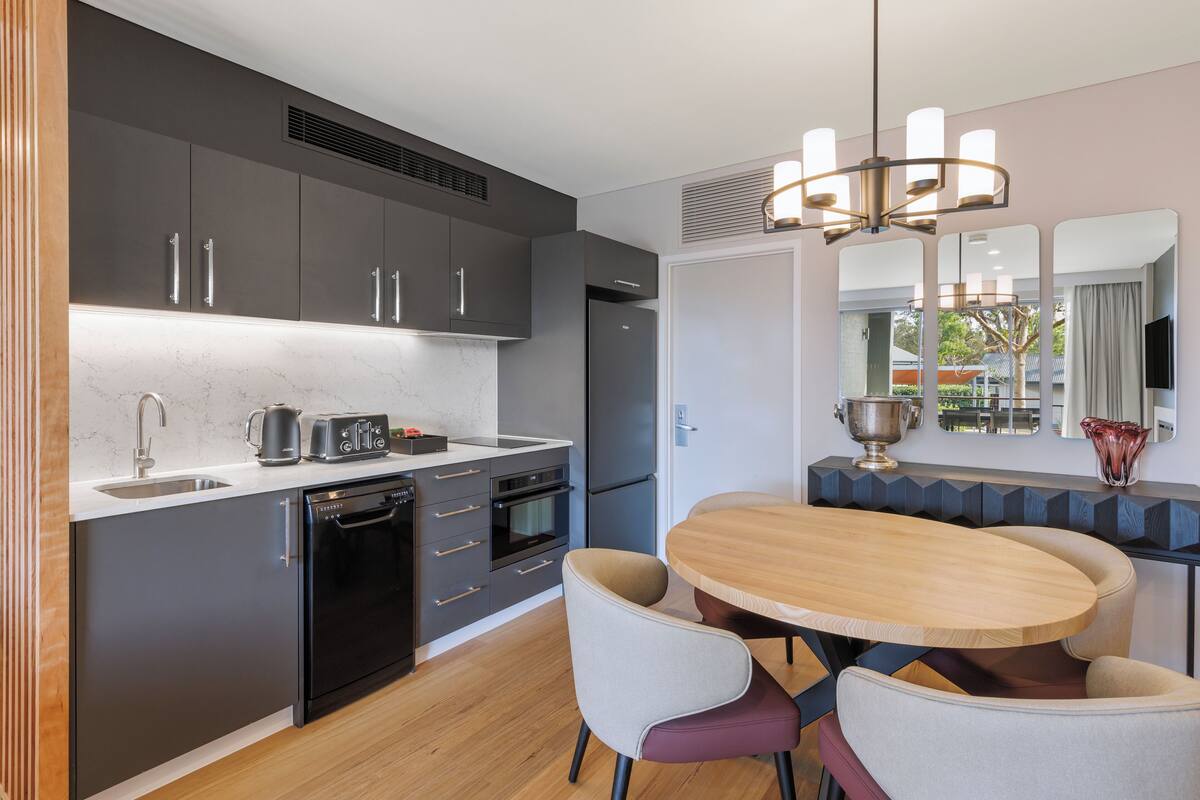 Una cocina moderna con armarios oscuros, horno empotrado y zona de comedor con mesa y sillas de madera.