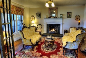 Una habitación con dos sillones tapizados en amarillo, una chimenea, un tapete rojo estampado y una mesa de madera con vasos y una botella.