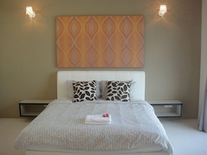 A neatly made bed with a patterned bedspread, two pillows with a leaf design, a bedside table, and a wall-mounted artwork above the bed. - Shamrock Beach Villas (George Town)