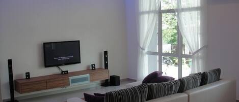 A modern living room with a flat-screen TV mounted on a wooden console, a large window with sheer curtains, and a white sofa with striped cushions.