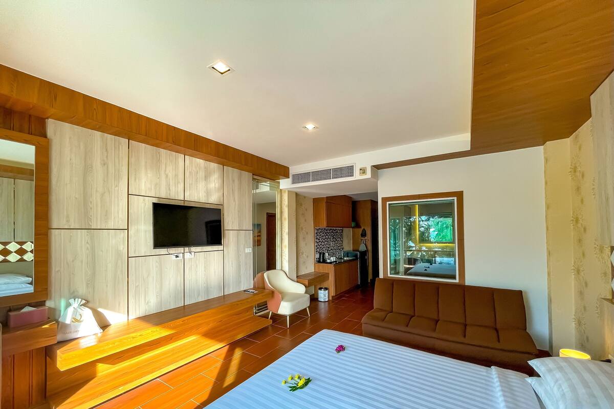 A modern hotel room with a large bed, a TV mounted on the wall, a wooden desk, and a seating area with a sofa and chair.
