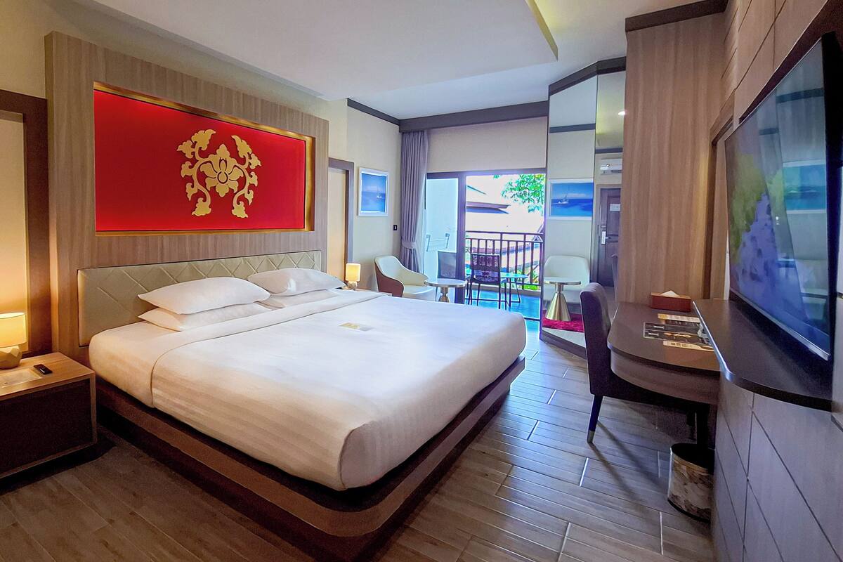 A hotel room with a large bed, a TV, a desk with a lamp, and a view of the outside balcony.