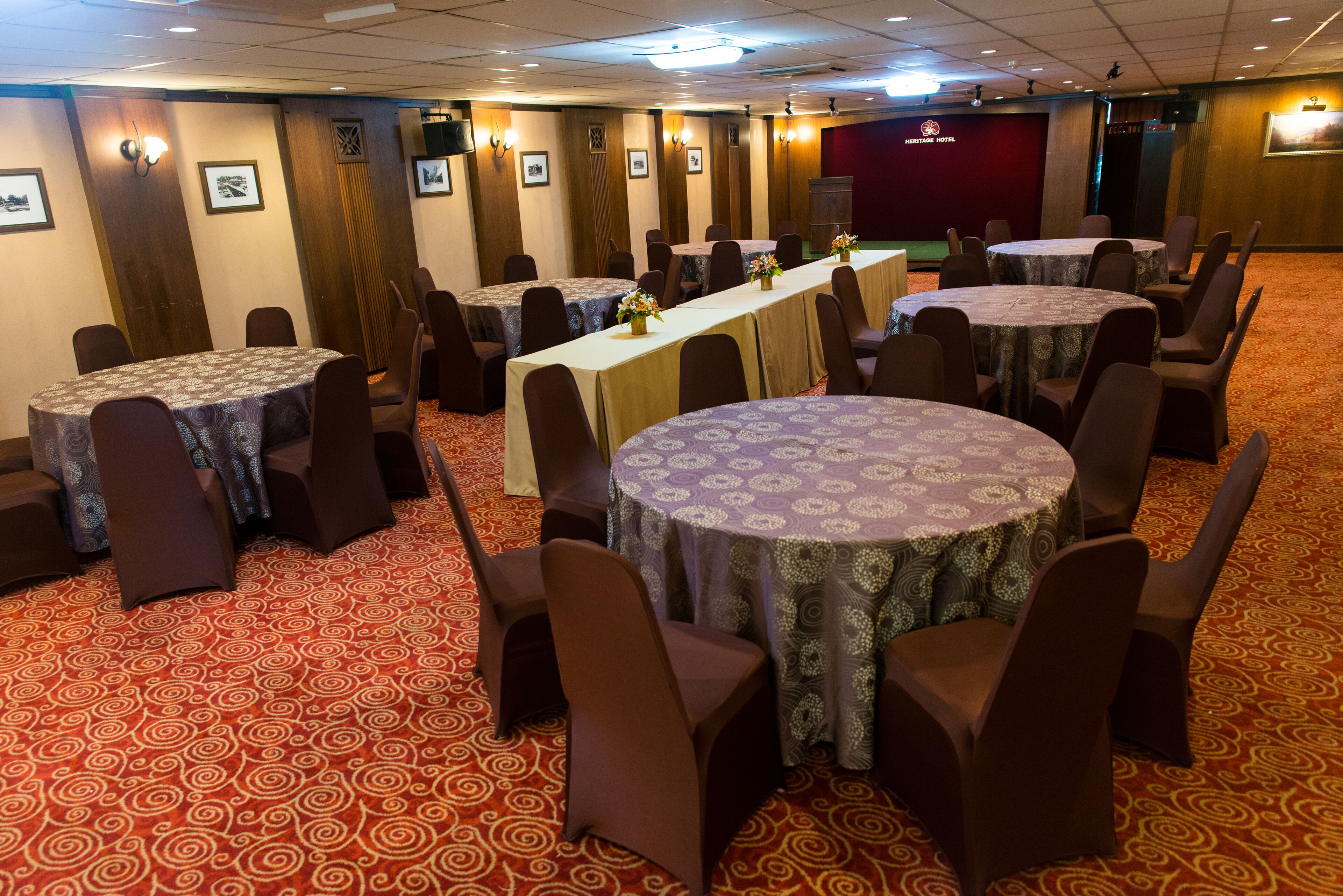 A conference room with round tables and chairs, a long table with a floral centerpiece, and a stage with a red backdrop.