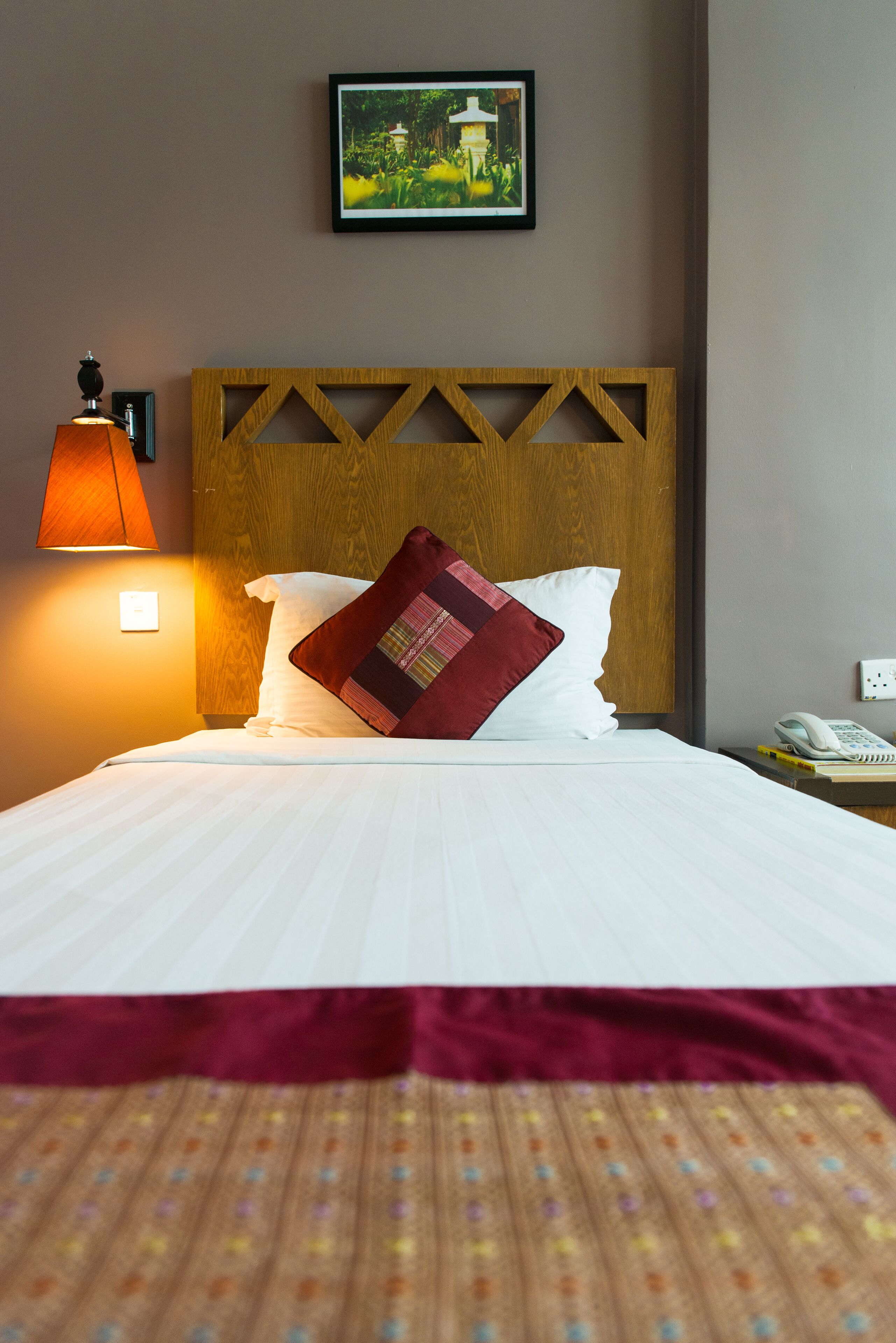 A neatly made bed with white linens, a wooden headboard, a red pillow, and a wall-mounted lamp.
