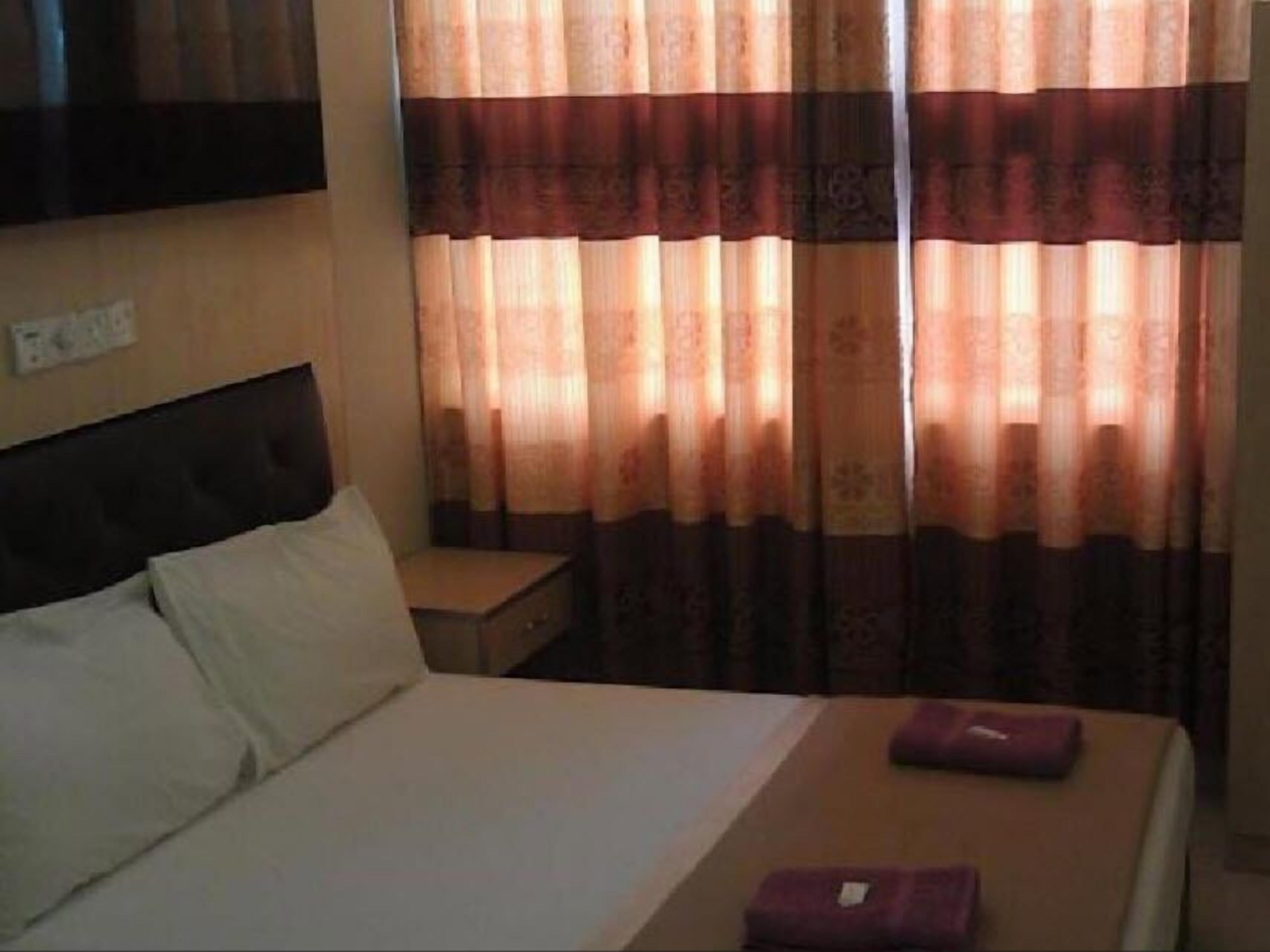 A hotel room with a bed, pillows, a nightstand, and a window with patterned curtains.