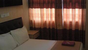 A hotel room with a bed, pillows, a nightstand, and a window with patterned curtains.