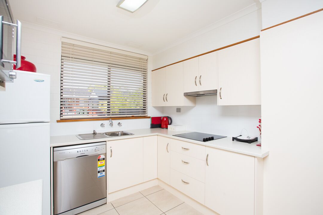 A modern kitchen with white cabinets, a stainless steel dishwasher, a built-in oven, a sink, and a window with blinds.