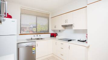 A modern kitchen with white cabinets, a stainless steel dishwasher, a built-in oven, a sink, and a window with blinds.