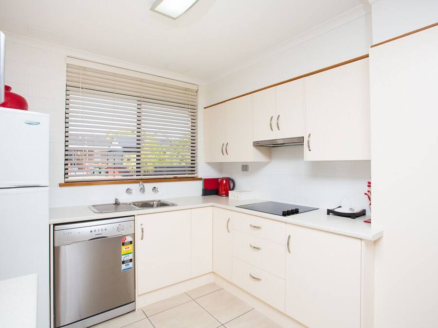 A modern kitchen with white cabinets, a stainless steel dishwasher, a built-in oven, a sink, and a window with blinds.