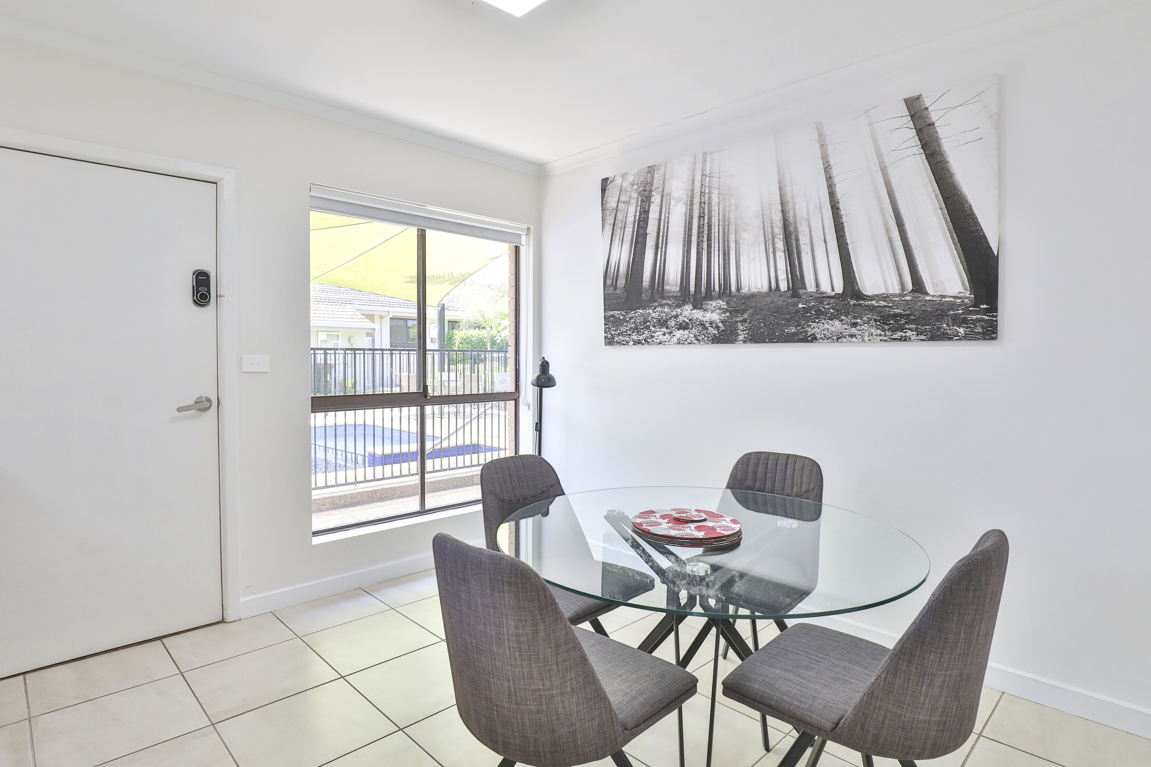 A dining area with a glass table and four chairs, a door with a keypad lock, and a large black and white forest painting on the wall.