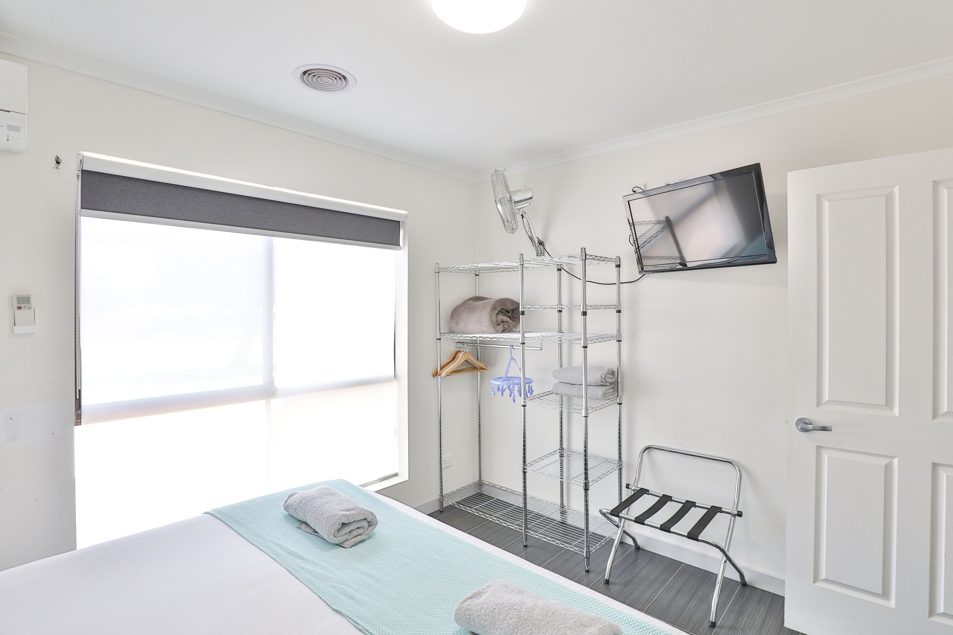 Habitación de hotel con cama, ventana con persiana, estantería metálica con toallas, una silla y televisor de pantalla plana en la pared.