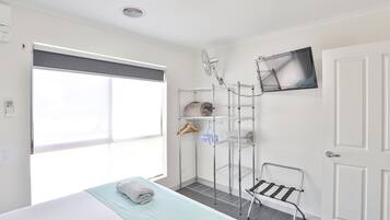 Habitación de hotel con cama, ventana con persiana, estantería metálica con toallas, una silla y televisor de pantalla plana en la pared.