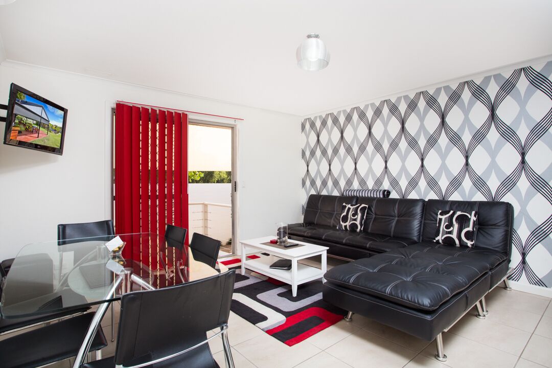 Una sala de estar moderna con un sofá de cuero negro, una mesa de comedor de vidrio con sillas, una mesa de centro blanca y una cortina roja.
