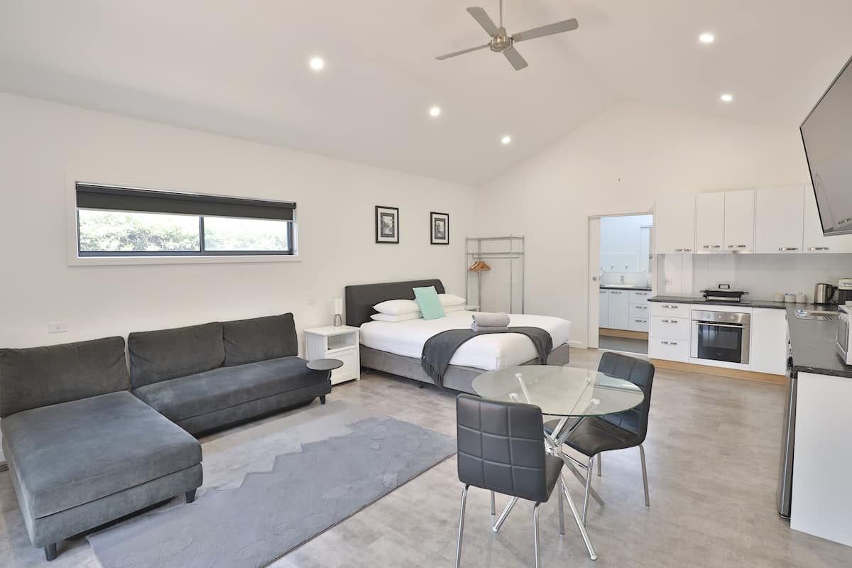 Un espace de vie moderne et compact comprenant un canapé, un lit, une table à manger avec des chaises, un coin cuisine et un ventilateur de plafond.