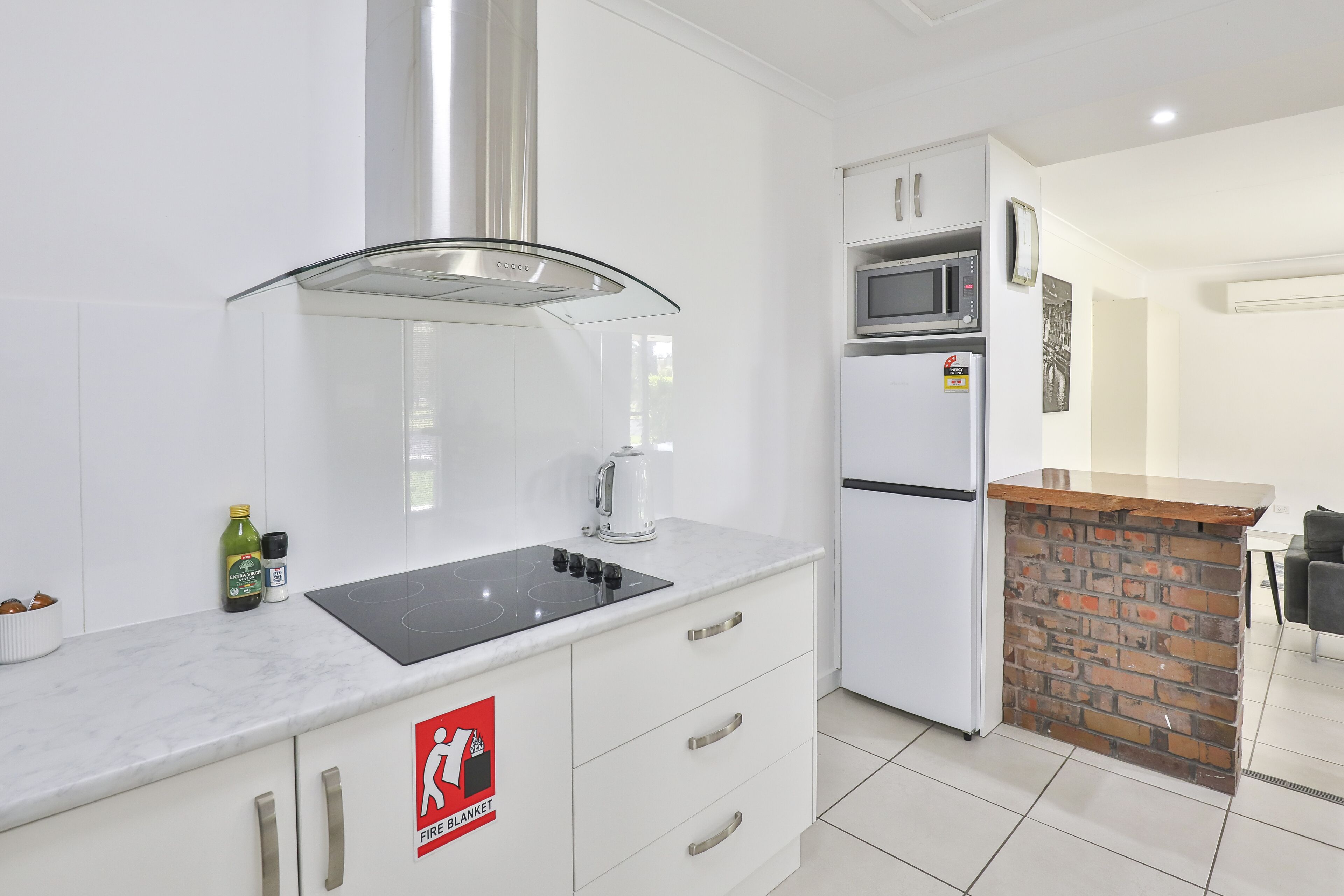 A modern kitchen with a white countertop, a stainless steel range hood, a microwave, and a refrigerator. There is a fire safety sign on the cabinet.