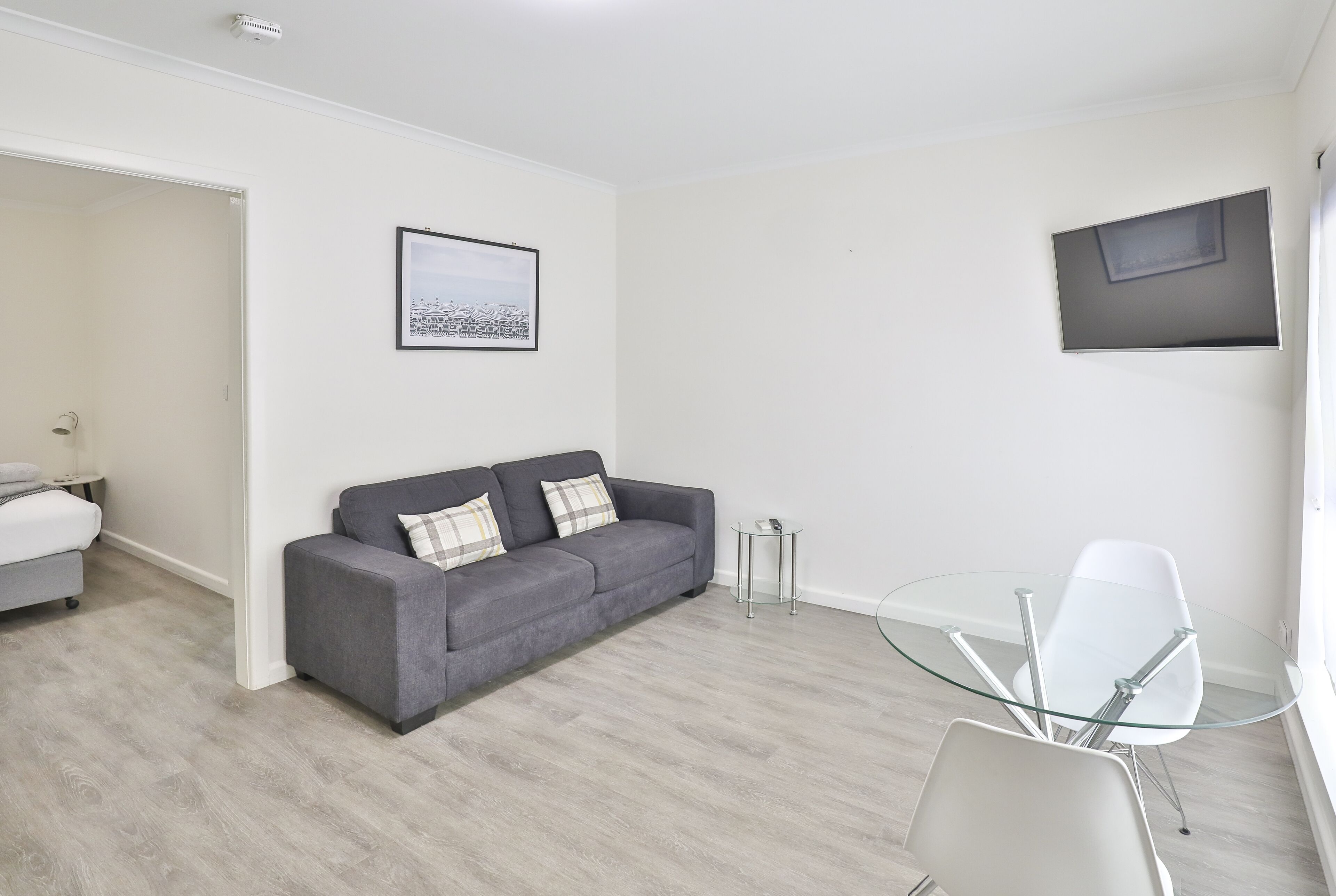 Un salón moderno con un sofá gris, una mesa de centro de vidrio y una silla blanca. Hay un cuadro en la pared y un televisor de pantalla plana montado sobre el sofá.
