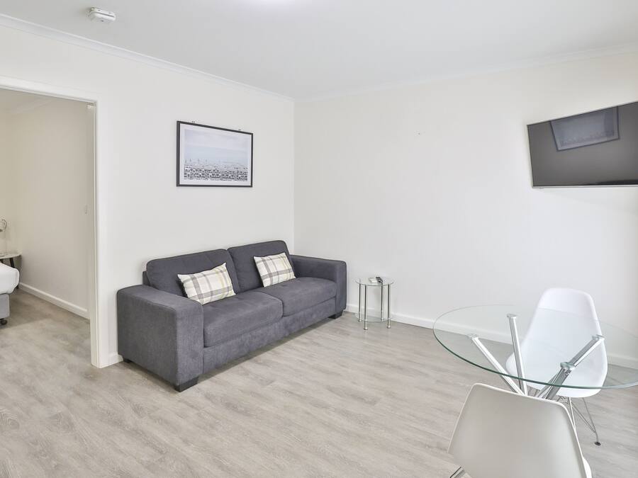 Un salón moderno con un sofá gris, una mesa de centro de vidrio y una silla blanca. Hay un cuadro en la pared y un televisor de pantalla plana montado sobre el sofá.