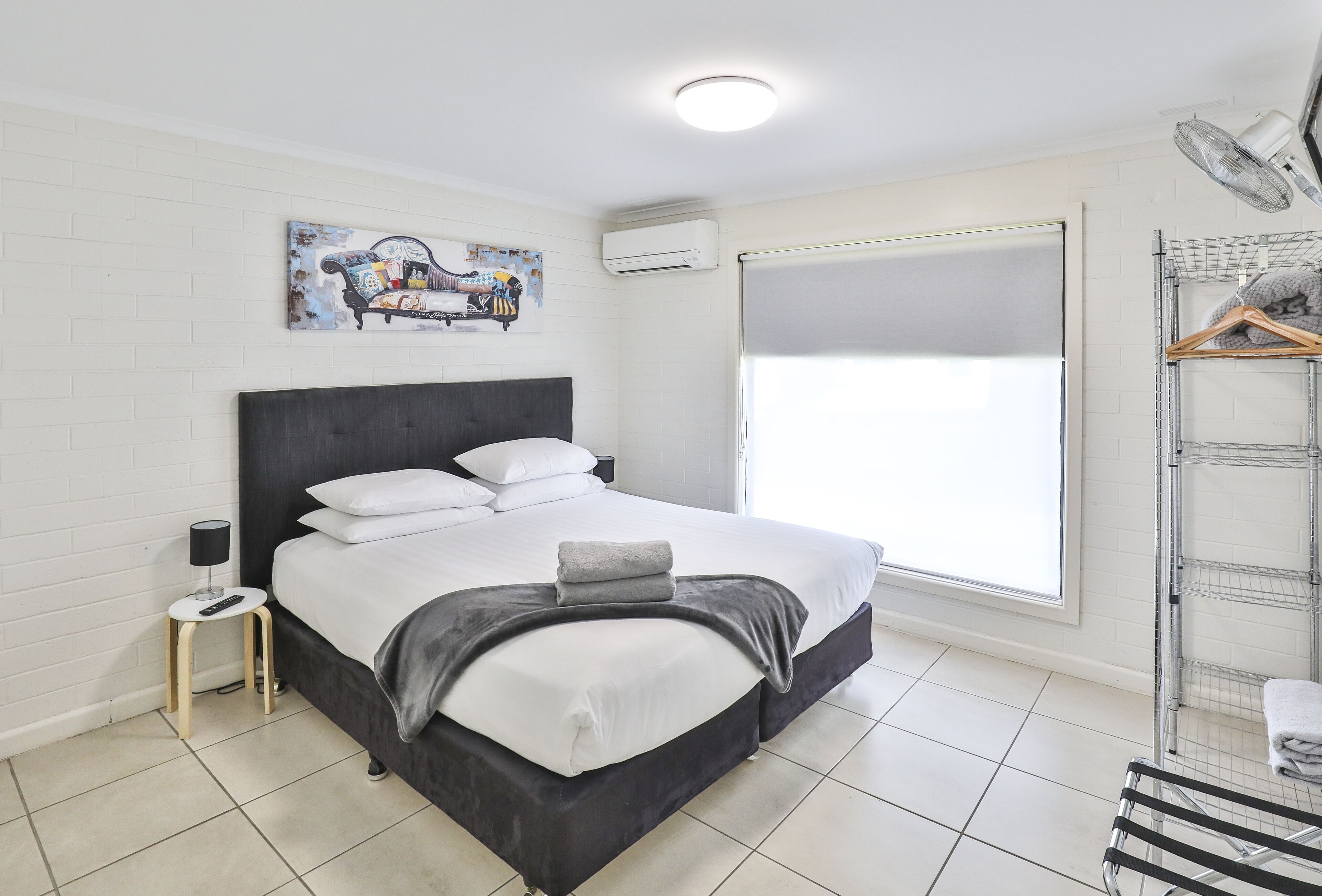 A modern hotel room with a large bed, a small side table, an air conditioning unit, a window with a view, and a laundry area with a drying rack.