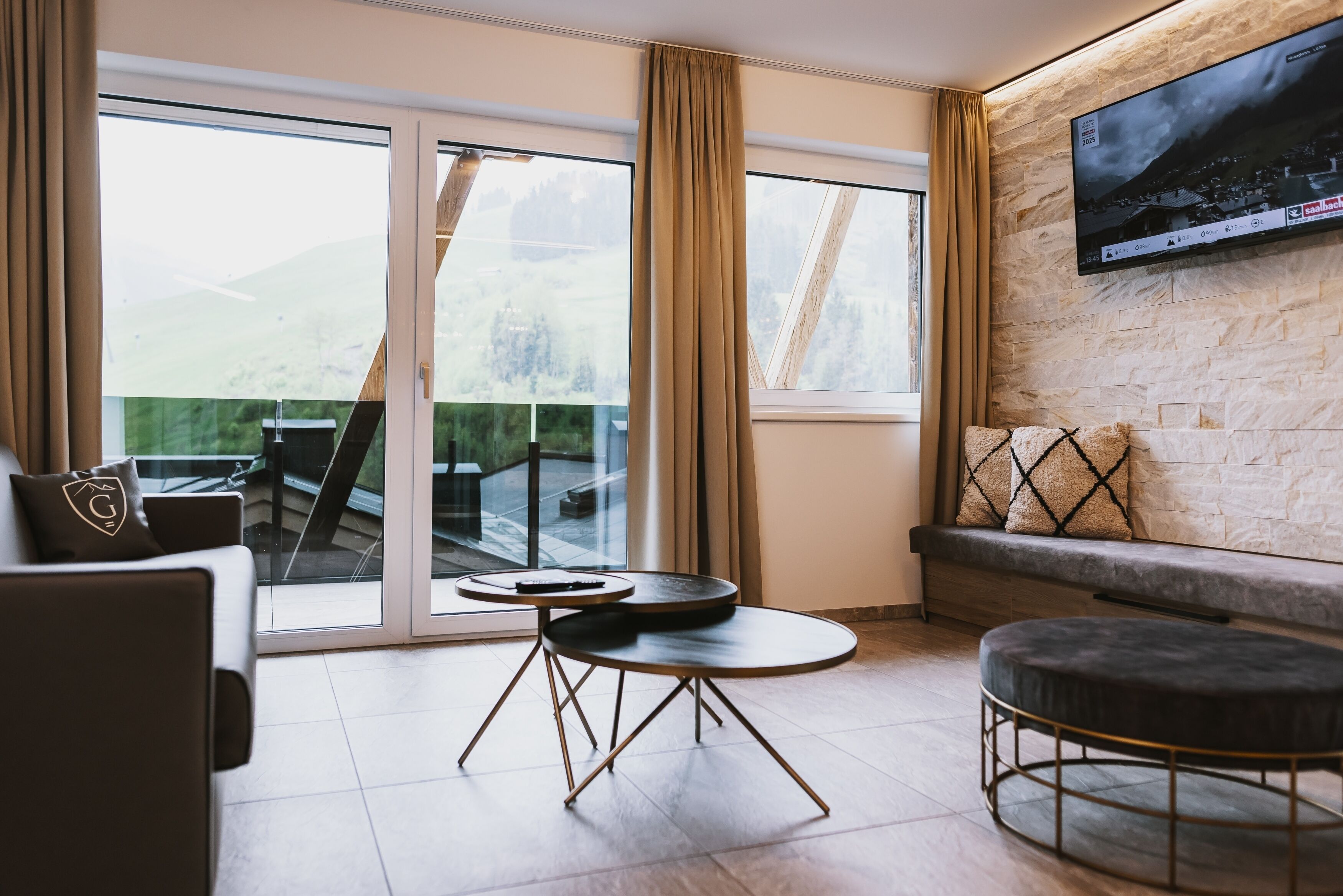 Un salon moderne doté d’une grande fenêtre, d’un téléviseur à écran plat fixé au mur et d’un coin salon confortable avec un canapé et un pouf.