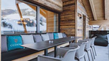 A wooden cabin interior with a long dining table, cushioned seating, and large windows offering a view of a snowy landscape.