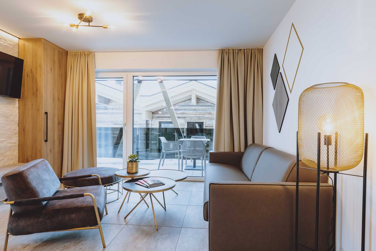 Ein modernes Wohnzimmer mit Sofa, Sesseln, Couchtisch und Stehlampe. Ein Fernseher ist an der Wand befestigt, und durch große Fenster ist ein Blick auf ein Gebäude möglich.