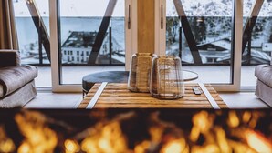 A cozy room with a fireplace, a wooden table with glass jars, and a view of snow-covered mountains through large windows. - GLEMM by AvenidA Superior Hotel & Residences (Saalbach-Hinterglemm)