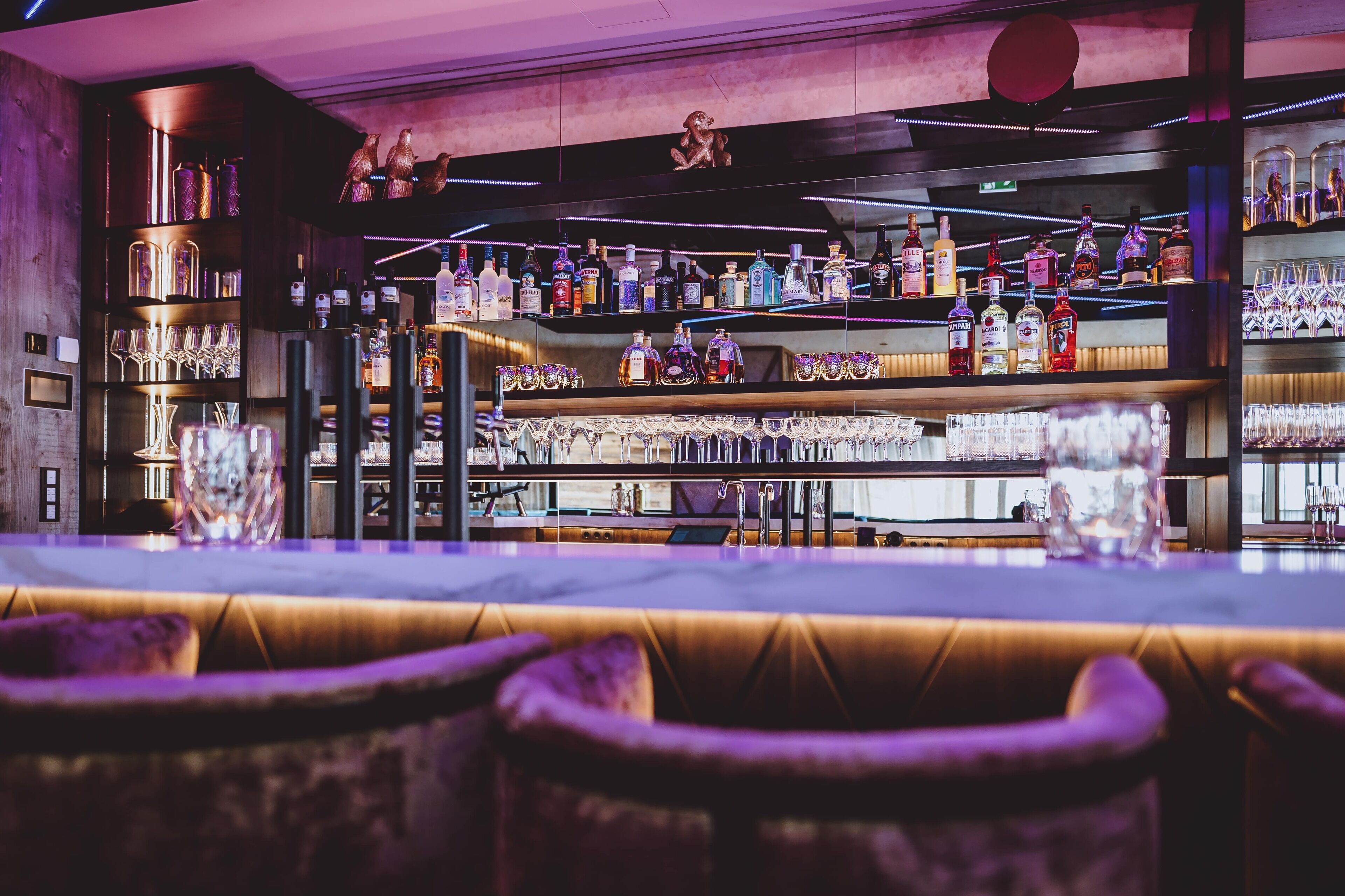 Zona de bar con varias estanterías llenas de botellas de licor y vasos, una barra y asientos acolchados.