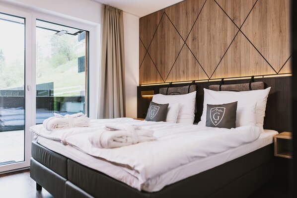 A neatly made bed with towels, a wooden headboard, and a view of the outdoors through a sliding door. - GLEMM by AvenidA Superior Hotel & Residences (Saalbach-Hinterglemm)
